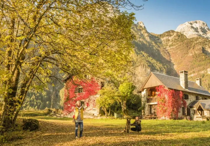 Dos mujeres y un perro disfrutan del otoño en el valle de Pineta, ideal para rutas. Las casas cubiertas de hiedra roja y las montañas crean un paisaje idílico.