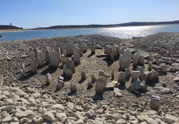El dolmen de Guadalperal emerge de las aguas bajas, un círculo de menhires ancestrales sobre un lecho de piedras y arena, con un lago sereno al fondo.