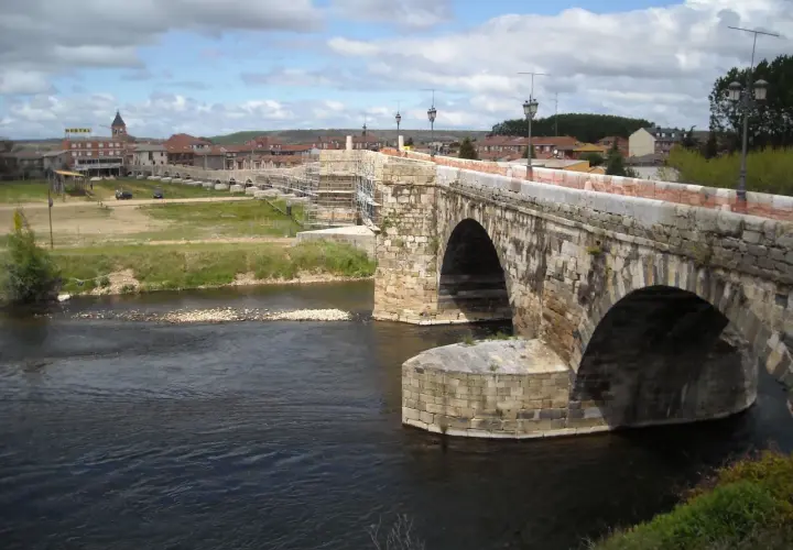 Puente romano en Hospital de Órbigo, con arcos de piedra y andamios. Al fondo, el pueblo y un hostal.