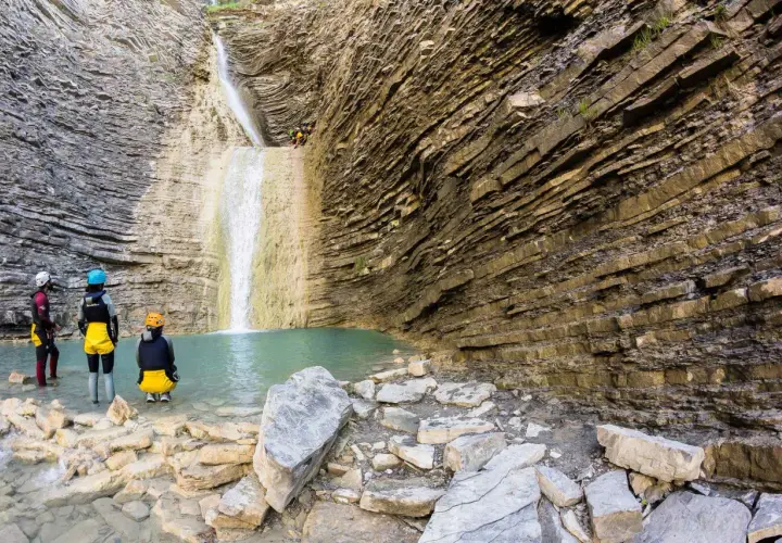 Un grupo de barranquistas se prepara para descender una cascada de oros bajo, rodeados de imponentes paredes rocosas estratificadas y una poza de agua turquesa.