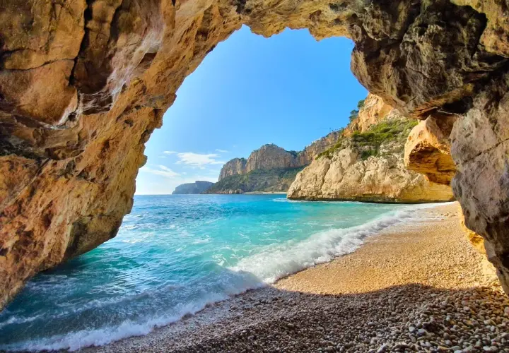 Vista desde una cueva en Cala Moraig, con aguas turquesas y acantilados dorados bajo un cielo azul.