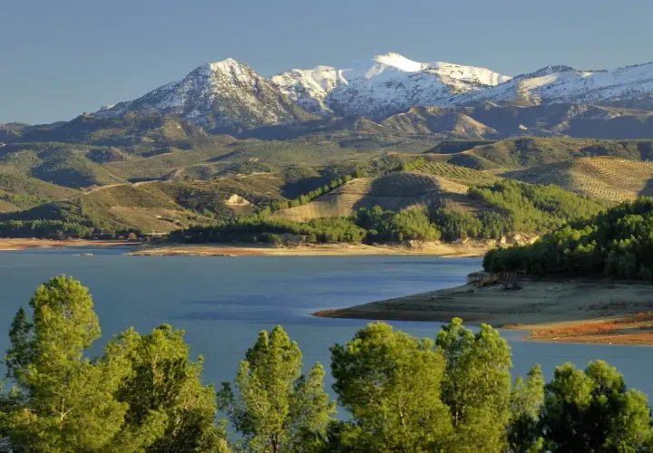 El pantano de los Bermejales, con montañas nevadas al fondo y colinas verdes salpicadas de olivos.