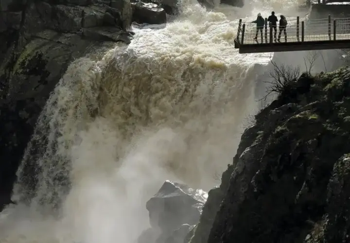 Tres personas observan la furia del pozo de los humos desde una pasarela. El agua turbia cae con fuerza, creando una densa niebla.