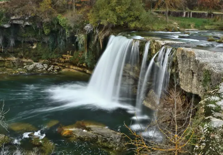La majestuosa cascada de Tobalina, con su velo de agua cayendo sobre rocas cubiertas de musgo, rodeada de árboles otoñales y un río sereno.