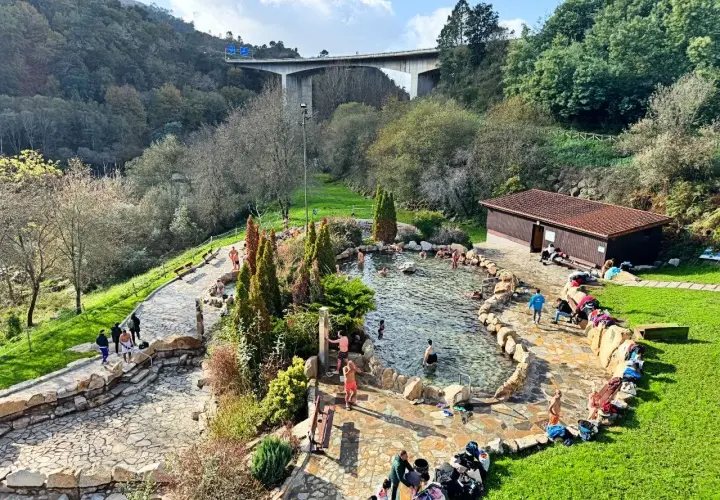Gente disfrutando de las **termas en Galicia**, un oasis de relajación rodeado de naturaleza, con un puente moderno al fondo.