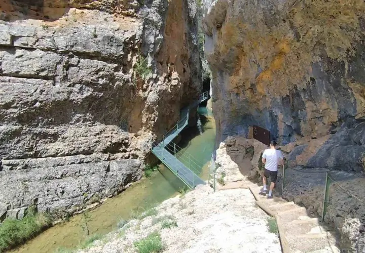 Un hombre camina por las pasarelas de Calomarde, un sendero metálico que serpentea sobre un río entre imponentes paredes rocosas.