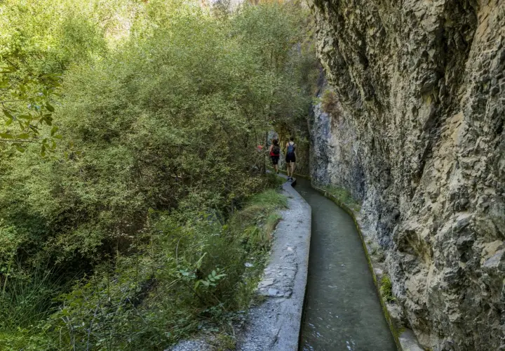Senderistas caminan por la ruta de los bolos, un sendero estrecho junto a un canal de agua y un acantilado rocoso.