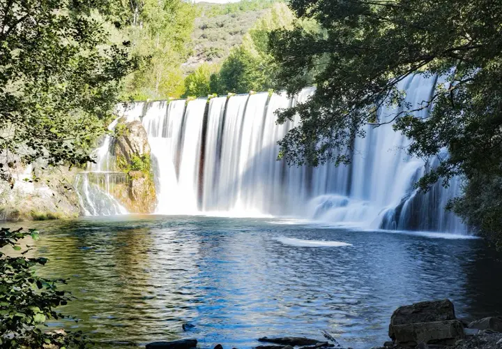 El Salto del Pelgo, una cascada imponente con agua sedosa cayendo sobre rocas, rodeada de vegetación exuberante y un estanque azul profundo.