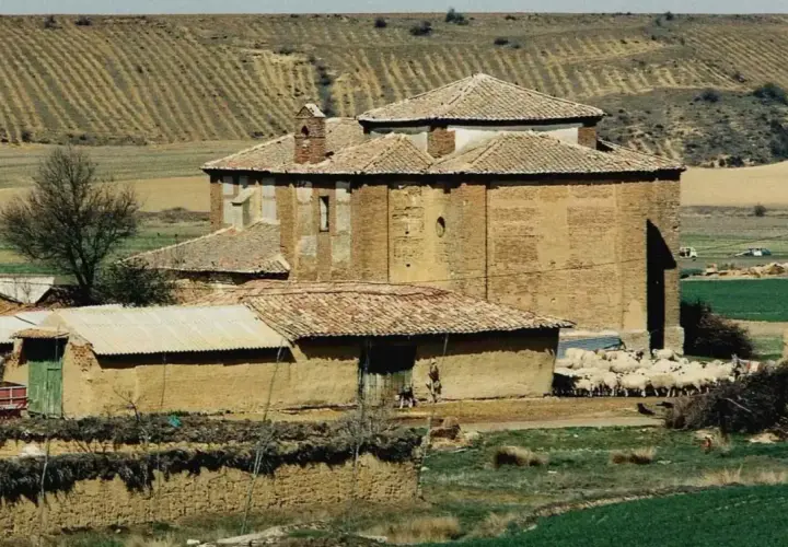 Edificio de ladrillo con tejado a dos aguas, rebaño de ovejas pastando cerca. Paisaje rural con campos de cultivo en terrazas, típico de calzadilla de la cueza.