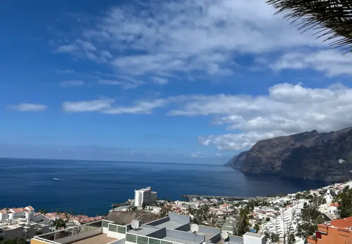 Vista panorámica de acantilados imponentes y el mar azul, con un pueblo costero y un puerto deportivo. Un lugar que evoca las mejores playas de Tenerife.