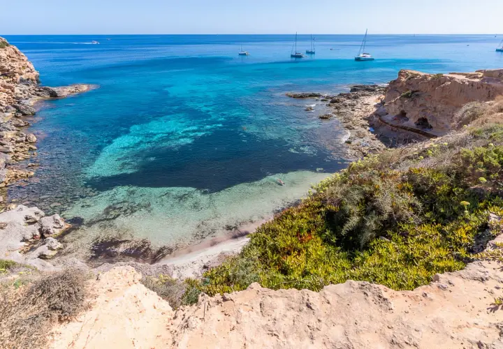 Aguas turquesas y arena blanca en una cala rocosa, un paraíso entre las mejores playas de Formentera. Yates navegan en el horizonte.