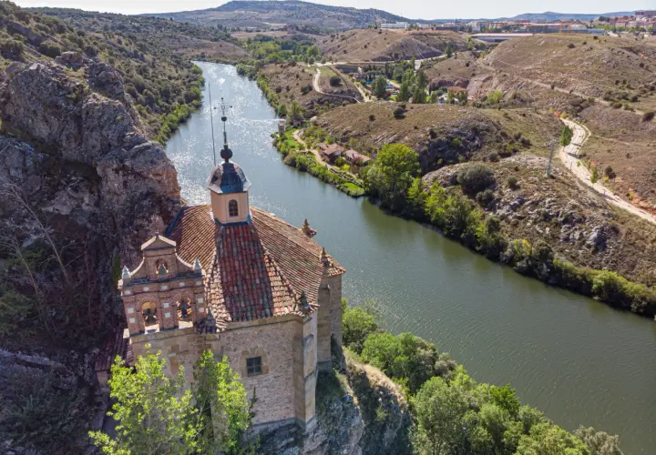 Ermita de San Saturio, Soria, se alza sobre un acantilado junto al río Duero, rodeada de vegetación y colinas.
