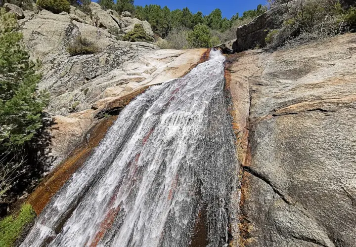 El agua desciende por la roca en la cascada del Hornillo, rodeada de vegetación y rocas graníticas bajo un cielo azul.