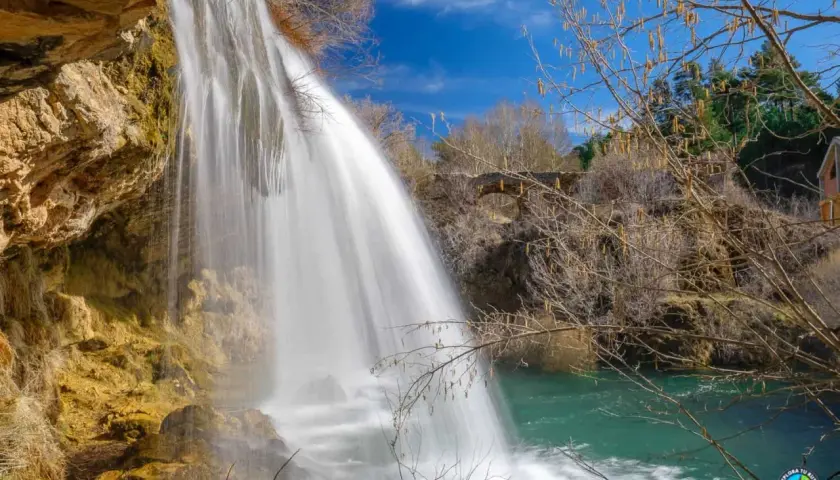 Cascada de agua blanca cayendo sobre rocas ocres, como los ojos del Cabriel, en un día azul.