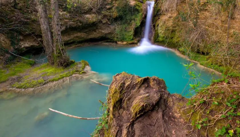 Cascada de agua turquesa en la ruta del agua Berganzo, rodeada de vegetación y rocas cubiertas de musgo.