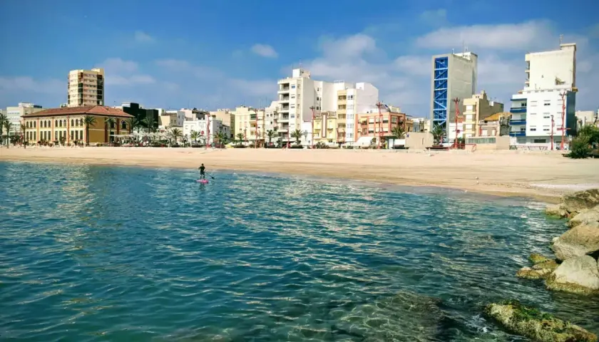 Un día soleado en la costa, con un paddleboarder solitario en el mar turquesa y edificios modernos de fondo. ¡Un oasis de playas cerca de Zaragoza!