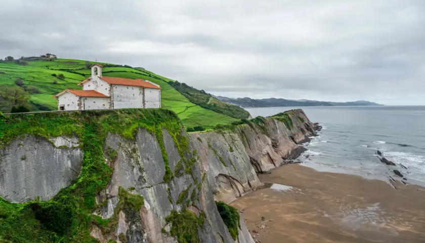 Iglesia blanca con tejado naranja sobre acantilado verde, junto al mar Cantábrico. Un lugar que evoca la esencia de "8 apellidos vascos".