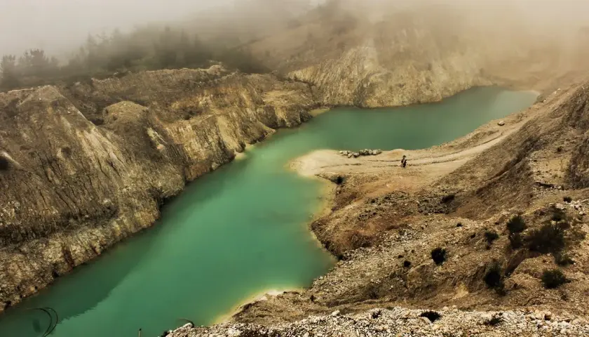 Laguna turquesa en el monte Neme Carballo, rodeada de tierra y rocas, con niebla al fondo.