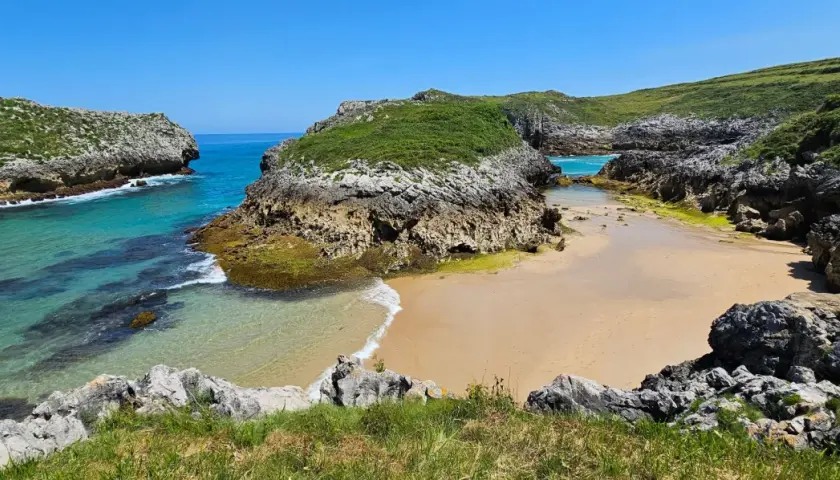 Una cala escondida entre acantilados verdes, con aguas turquesas y arena dorada. Un rincón idílico entre las mejores playas de Asturias.