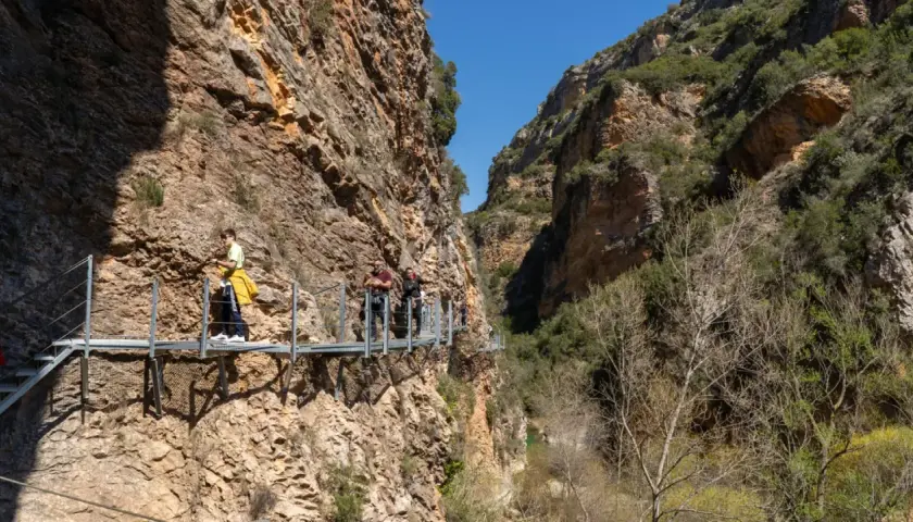Gente camina por las pasarelas de Alquézar, un sendero suspendido entre imponentes paredes rocosas y vegetación.