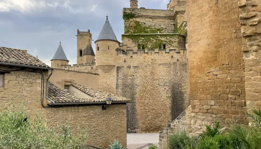 El **castillo de Olite**, con sus torres cónicas y muros de piedra, se alza majestuoso bajo un cielo nublado. En primer plano, tejados de tejas y vegetación verde añaden un toque de vida.