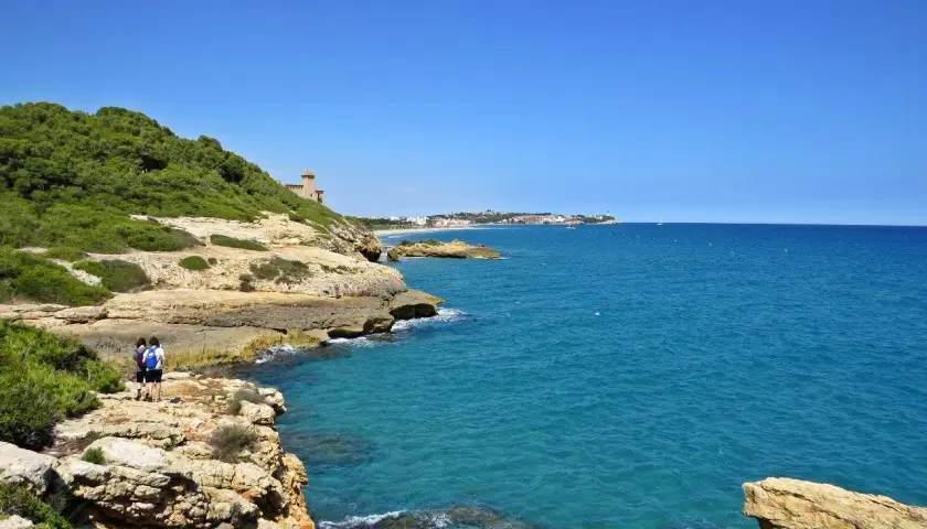 Dos personas caminan por el camí de ronda de Tarragona, disfrutando de las vistas al mar azul y la costa rocosa.