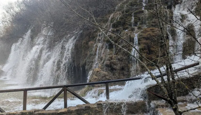 Cascada en la ruta nacimiento río Cuervo. Agua cayendo sobre rocas cubiertas de musgo y vegetación. Barandilla de madera en primer plano.