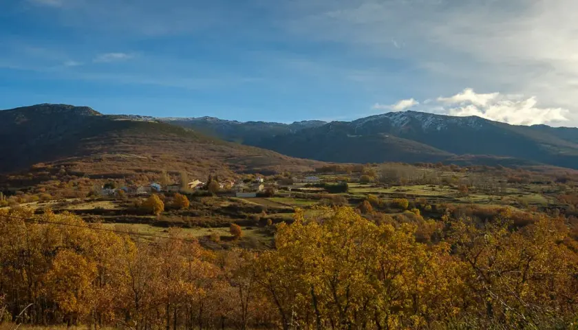 Paisaje otoñal con un pueblo en la ladera. Las montañas nevadas al fondo sugieren la ruta a la laguna negra.