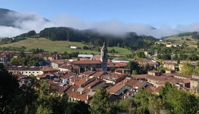 Vista panorámica de Oñati, con su iglesia y tejados rojizos, rodeada de verdes colinas y niebla. Ideal para planificar cómo llegar.