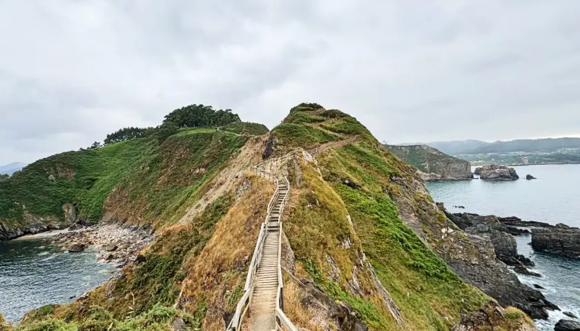 Un sendero de madera asciende por una colina verde, con el mar azul a la derecha. La forma de la costa recuerda al fuciño do porco.