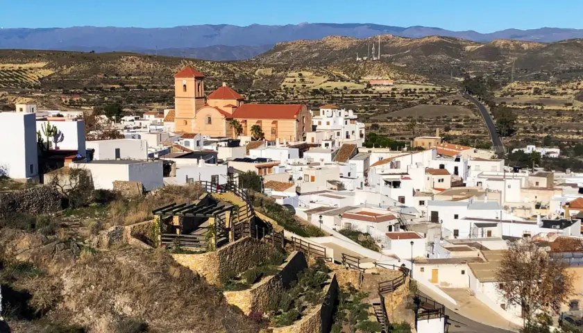 Vista panorámica de Lucainena de las Torres, un pueblo blanco andaluz con una iglesia de campanario naranja. El camino para llegar serpentea entre colinas áridas.