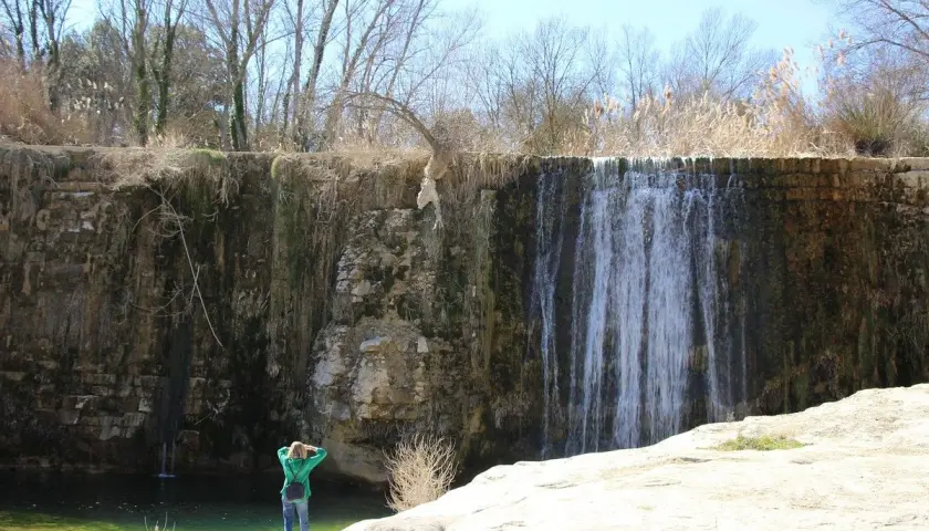 Una persona admira una cascada en un paisaje rocoso. El agua cae sobre la pared de roca, creando un velo. El sol brilla, y la vegetación seca rodea la escena. Pozan de Vero, un lugar de belleza natural.