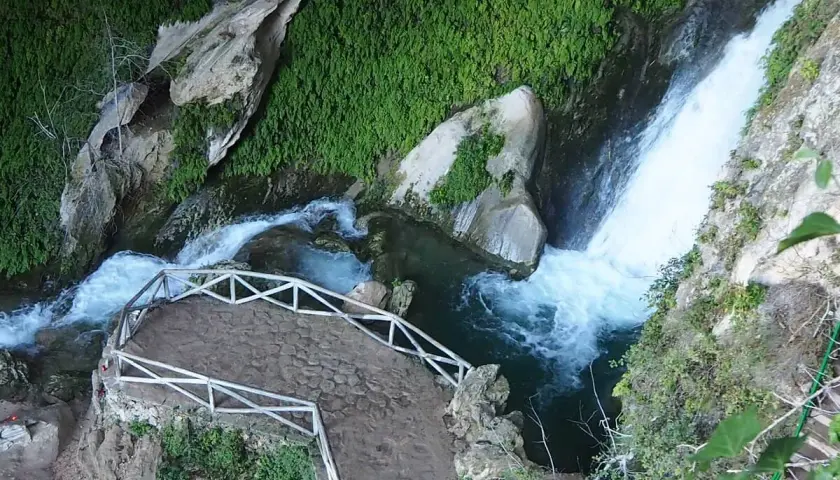 Sendero de piedra y barandilla blanca junto a la cascada de la cueva del agua Tiscar, rodeada de vegetación exuberante.