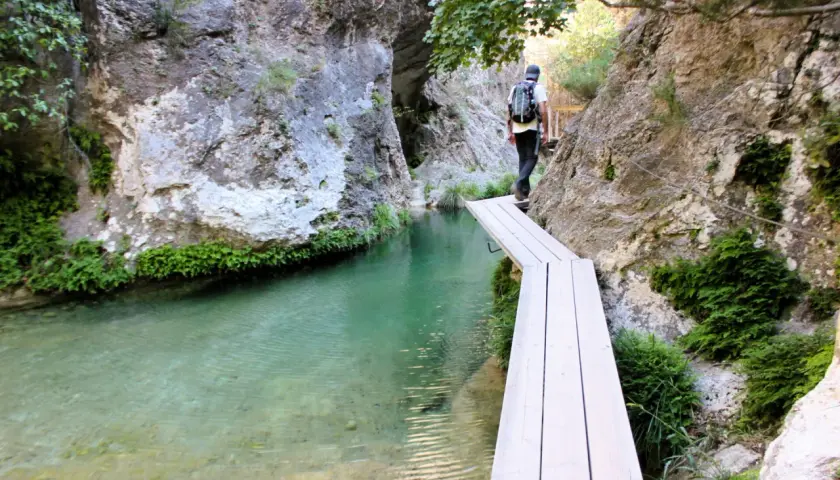 Senderista cruza un puente de madera sobre el agua cristalina en el Parrizal de Beceite, rodeado de rocas y vegetación exuberante.