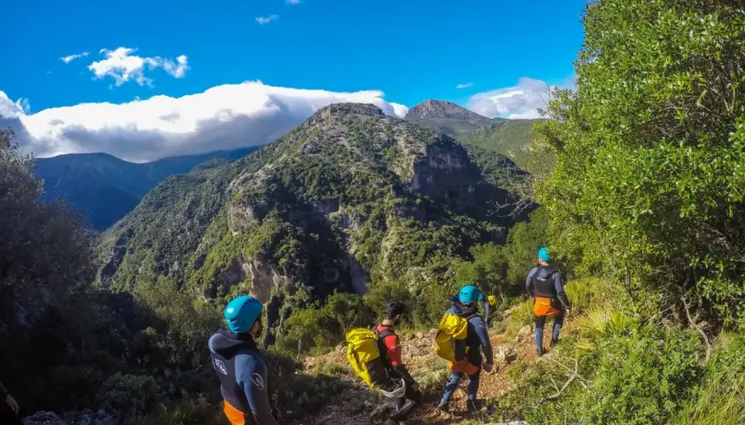 Grupo de barranquistas desciende por un sendero rocoso hacia la garganta verde, rodeados de exuberante vegetación y montañas.