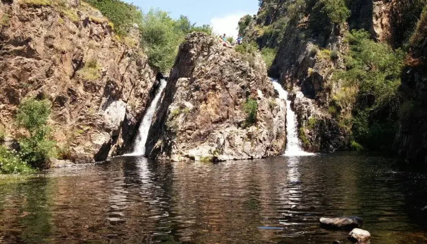 Dos cascadas caen por rocas escarpadas en la cascada del hervidero, formando un estanque oscuro. Personas observan desde lo alto.