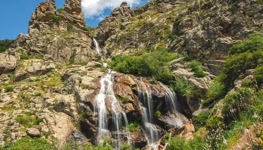 La cascada de los litueros desciende por rocas escarpadas, rodeada de vegetación exuberante bajo un cielo azul con nubes.