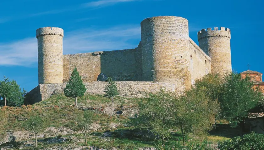 El castillo de Cornago se alza imponente sobre una colina, con sus torres circulares y murallas de piedra bajo un cielo azul intenso.
