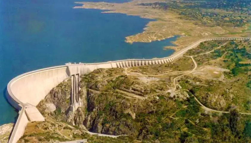 Vista aérea de la imponente presa de Almendra, reteniendo un vasto embalse de agua azul. La estructura curva se alza majestuosa sobre un paisaje rocoso y verde.