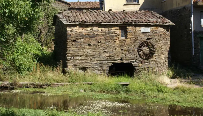 Antiguo molino de agua de piedra, con su rueda oxidada y vegetación exuberante a su alrededor.