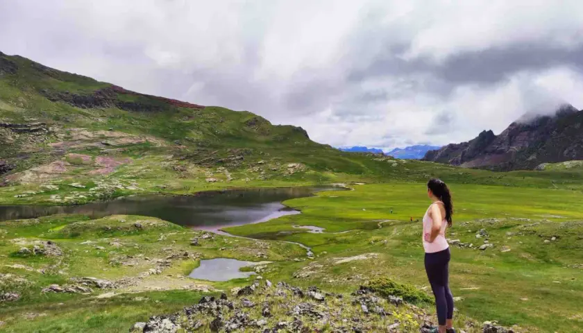 Mujer contempla los ibones de Anayet, lagos de montaña rodeados de verdes prados y picos rocosos bajo un cielo nublado.
