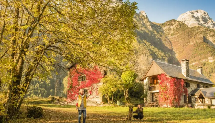 Dos mujeres y un perro disfrutan del otoño en el valle de Pineta, ideal para rutas. Las casas cubiertas de hiedra roja y las montañas crean un paisaje idílico.