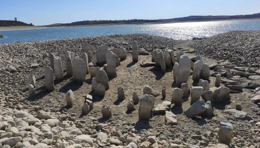 El dolmen de Guadalperal emerge de las aguas bajas, un círculo de menhires ancestrales sobre un lecho de piedras y arena, con un lago sereno al fondo.