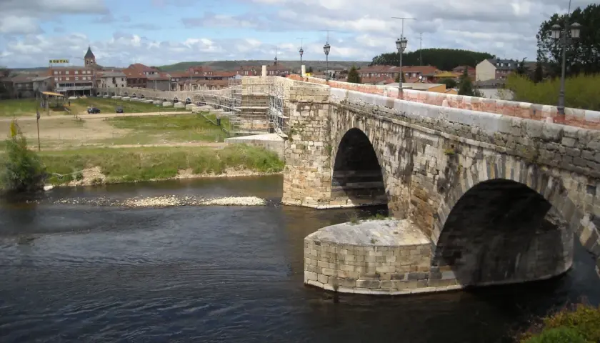 Puente romano en Hospital de Órbigo, con arcos de piedra y andamios. Al fondo, el pueblo y un hostal.
