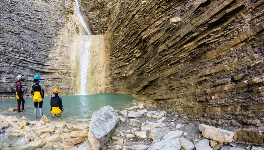 Un grupo de barranquistas se prepara para descender una cascada de oros bajo, rodeados de imponentes paredes rocosas estratificadas y una poza de agua turquesa.