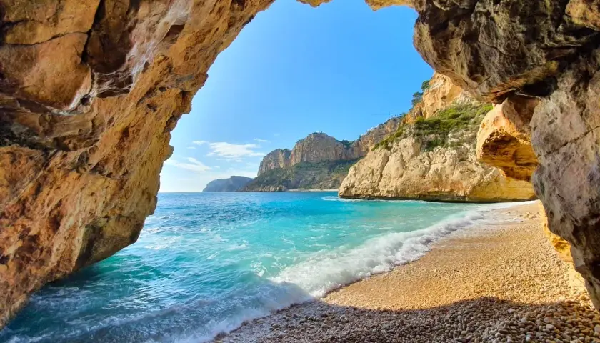 Vista desde una cueva en Cala Moraig, con aguas turquesas y acantilados dorados bajo un cielo azul.