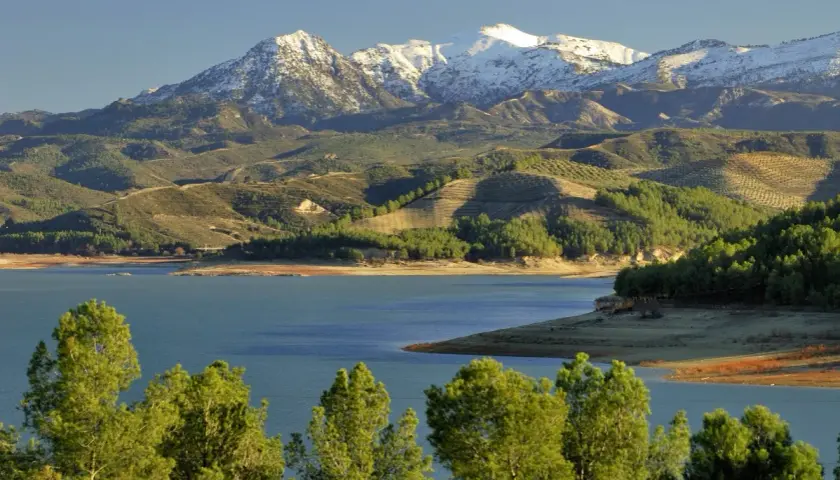 El pantano de los Bermejales, con montañas nevadas al fondo y colinas verdes salpicadas de olivos.