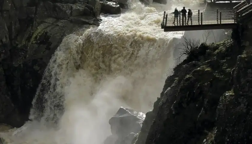 Tres personas observan la furia del pozo de los humos desde una pasarela. El agua turbia cae con fuerza, creando una densa niebla.