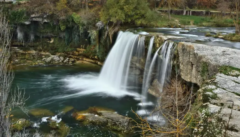 La majestuosa cascada de Tobalina, con su velo de agua cayendo sobre rocas cubiertas de musgo, rodeada de árboles otoñales y un río sereno.