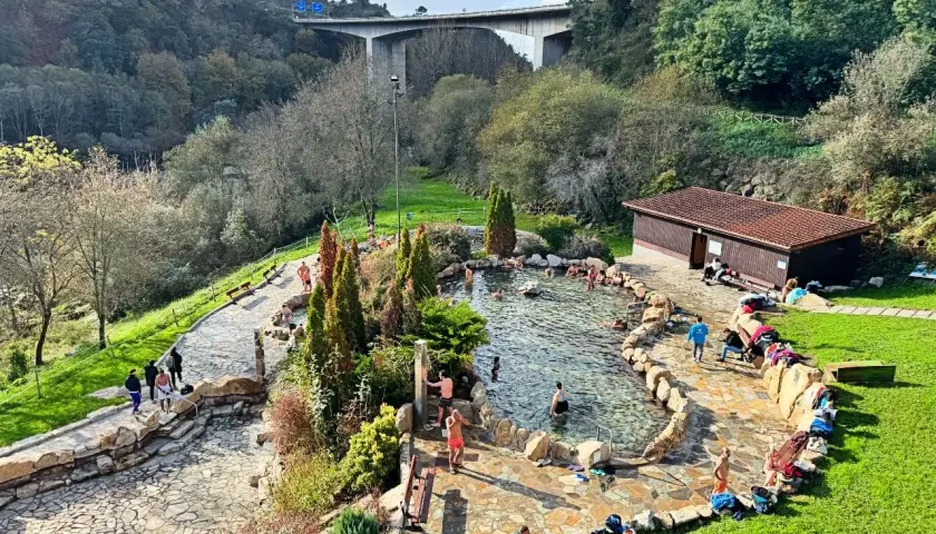 Gente disfrutando de las **termas en Galicia**, un oasis de relajación rodeado de naturaleza, con un puente moderno al fondo.